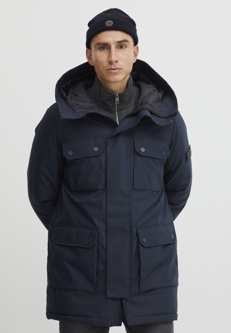 Marineblå isoleret parka med store frontlommer, hætte og lynlås foran. Har et blødt inderslag og en knapdetalje på ærmet.