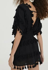 Vestido negro con tejido fluido, mangas cortas y cintura fruncida. Presenta capas texturizadas y detalles de borlas en el dobladillo y las mangas.