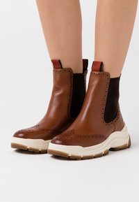 Botines de cuero marrón con paneles laterales de malla, suela blanca y perforaciones decorativas. Cuenta con lengüetas elásticas para un fácil calzado.