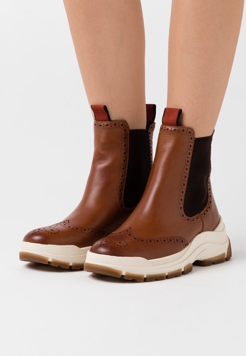 Botines de cuero marrón con paneles laterales de malla, suela blanca y perforaciones decorativas. Cuenta con lengüetas elásticas para un fácil calzado.