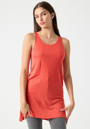 Mujer joven que lleva una túnica coral sin mangas y jeans grises, de pie contra un fondo claro y sencillo.