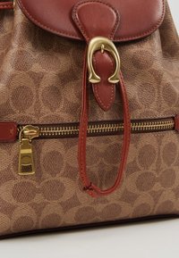 Zaino in tessuto a motivi marroni con flap in pelle rossa, accessori in oro, zip e un gancio decorativo. Progettato per funzionalità e stile.