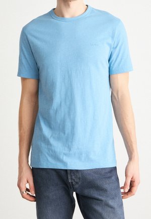 Mies, joka pukeutuneena yksinkertaiseen vaaleansiniseen lyhythihaiseen T-paitaan ja tummansinisiin farkkuihin, kädessään vasemmassa kädessä hopeasormus.