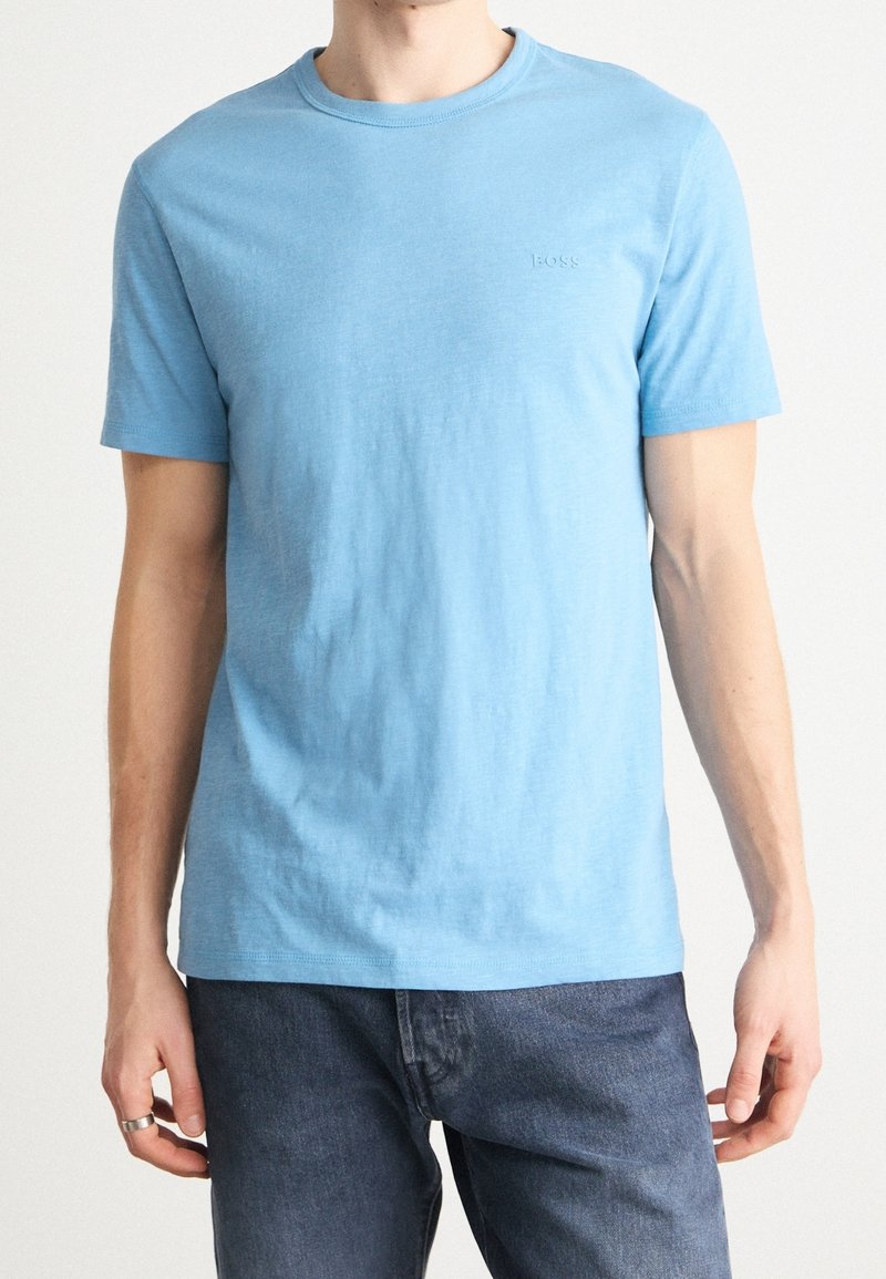 Homme portant un t-shirt à manches courtes uni bleu clair et un jean bleu foncé, avec une bague en argent à la main gauche.