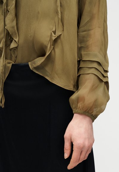 Ελαφριά μπλούζα σε ελιά πράσινο με βολάν και στρωμένα μανίκια με φουσκώματα, συνδυασμένη με μαύρη φούστα, με το χέρι χαλαρό στο πλάι.