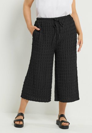 Person iført teksturerede, sorte vide cropped bukser med snøre i taljen og sorte platformsandaler mod en ensfarvet baggrund.