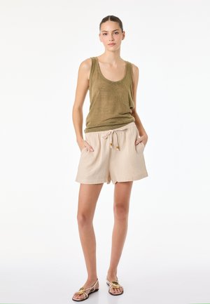 Ung kvinde stående med hænderne i lommerne, iført olivengrøn tanktop, beige shorts med snøre og beige sandaler med sløjfer.