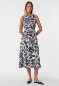 Ärmelloses Blumenkleid in Hellblau mit dunkelblauen und weißen Mustern. Mittel-lang, mit taillierter Taille, fließendem Rock, kombiniert mit schwarzen Schuhen.