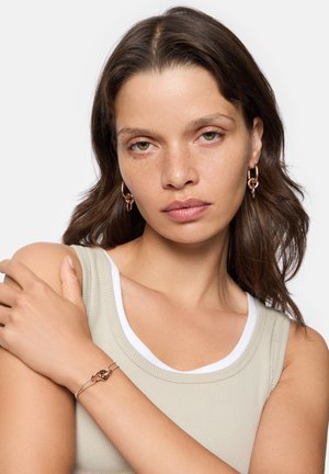 Des boucles d'oreilles en or rose en forme de créole et un bracelet relié. Le modèle porte un débardeur côtelé vert clair avec une bande blanche. La peau a une finition naturelle.