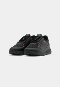 Black athletic shoes with mesh panels, red accents, and thick, textured soles. Features laces and branding on the tongue and side.