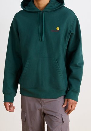 Mann trägt dunkelgrünes Carhartt Kapuzensweatshirt und hellgraue Cargohosen, steht vor einem schlichten Hintergrund.