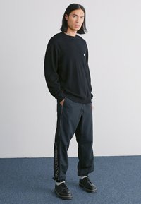 Black long-sleeve sweatshirt with a small logo, paired with black, tapered track pants featuring a side stripe and black lace-up shoes.