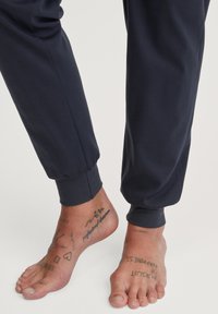 Marine joggers met elastische boorden en een gladde textuur, gecombineerd met blote voeten die tatoeages op de enkel en voet tonen.
