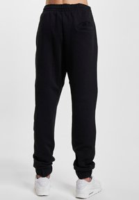 Schwarze Jogginghose mit elastischem Bund, Seitentaschen und gerippten Säumen. Weicher Stoff mit glatter Textur. Getragen mit weißen Turnschuhen.