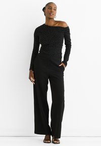 Black long-sleeve top with off-shoulder design, textured fabric featuring white dots. Paired with wide-legged black trousers and strappy sandals.