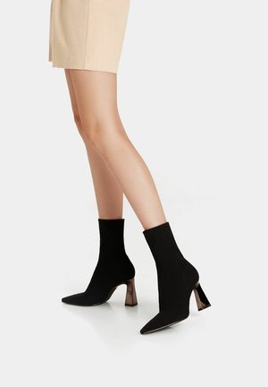 Bottes noires à la cheville en tissu doux avec un design à bout pointu et un talon métallique épais, textures contrastées, assorties à une jupe beige clair.