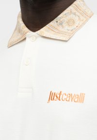 Polo blanc avec un col orné d'un motif beige et un logo "Just Cavalli" orange brodé sur la poitrine.