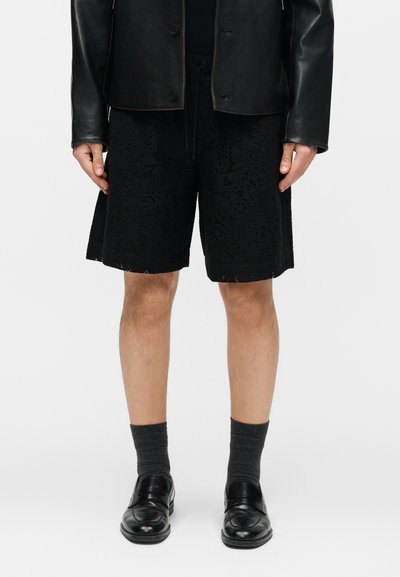 Άνδρας που φοράει μαύρα δαντελωτά σορτς μέχρι το γόνατο, μαύρο δερμάτινο μπουφάν, σκουρόχρωμες γκρι κάλτσες και μαύρα δερμάτινα μοκασίνια σε απλό φόντο.