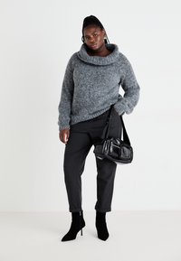 Suéter gris grueso de punto con un amplio cuello estilo cowl, combinado con pantalones negros a medida de cintura alta y botas de tobillo negras con puntera. Bolsa de mano negra.