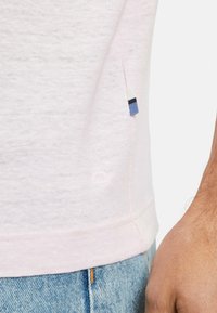 T-shirt in cotone rosa chiaro con una fessura laterale, caratterizzato da un'etichetta di accento blu navy e blu all'orlo. La texture è morbida e leggermente melangiata.