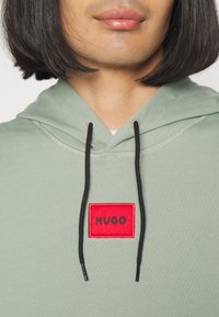Felpa verde chiaro con cappuccio e cordino, dotata di una patch rettangolare rossa con la scritta "HUGO" in testo nero. Tessuto morbido, design casual.