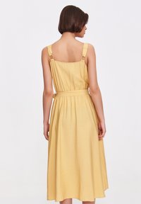 Żółta sukienka z regulowanymi ramiączkami i okrągłym okuciem. Posiada dopasowaną górę, paskowaną talię oraz zwiewną spódnicę o długości do kolan. Gładka tekstura.