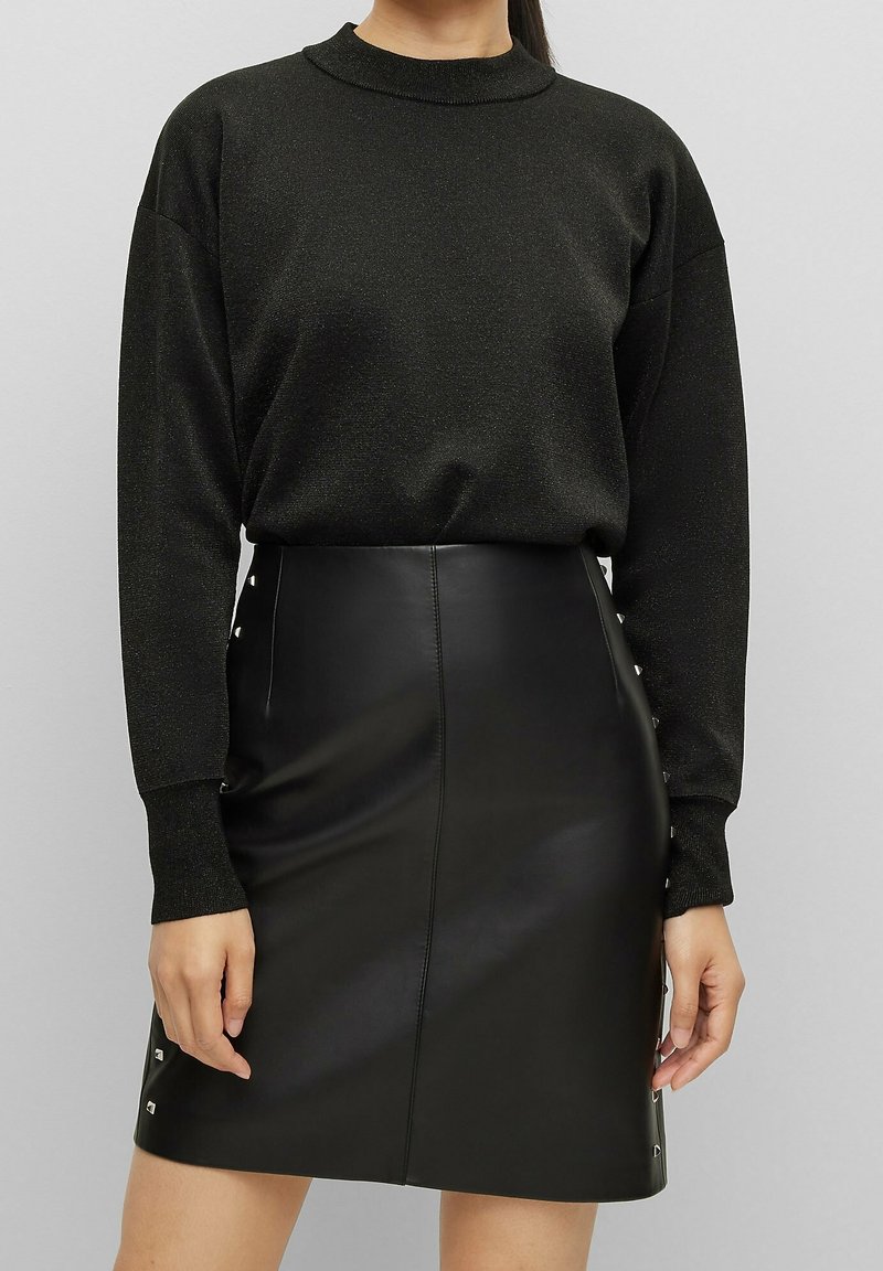 Maglione nero a maglia con collo alto e maniche lunghe, abbinato a una mini gonna in pelle nera con dettagli a bottoni a pressione sui lati.