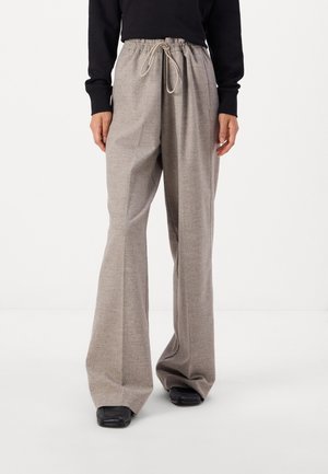 Osoba nosząca luźne, beżowe spodnie z szerokimi nogawkami i sznurkiem ściągającym oraz czarne buty, stojąca na tle jednolitego białego tła.