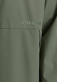 Zöld szövetdzseki sima textúrával, elöl kiemelkedő "JACK & JONES" logóval. Minimalista dizájn, további díszítések nélkül.