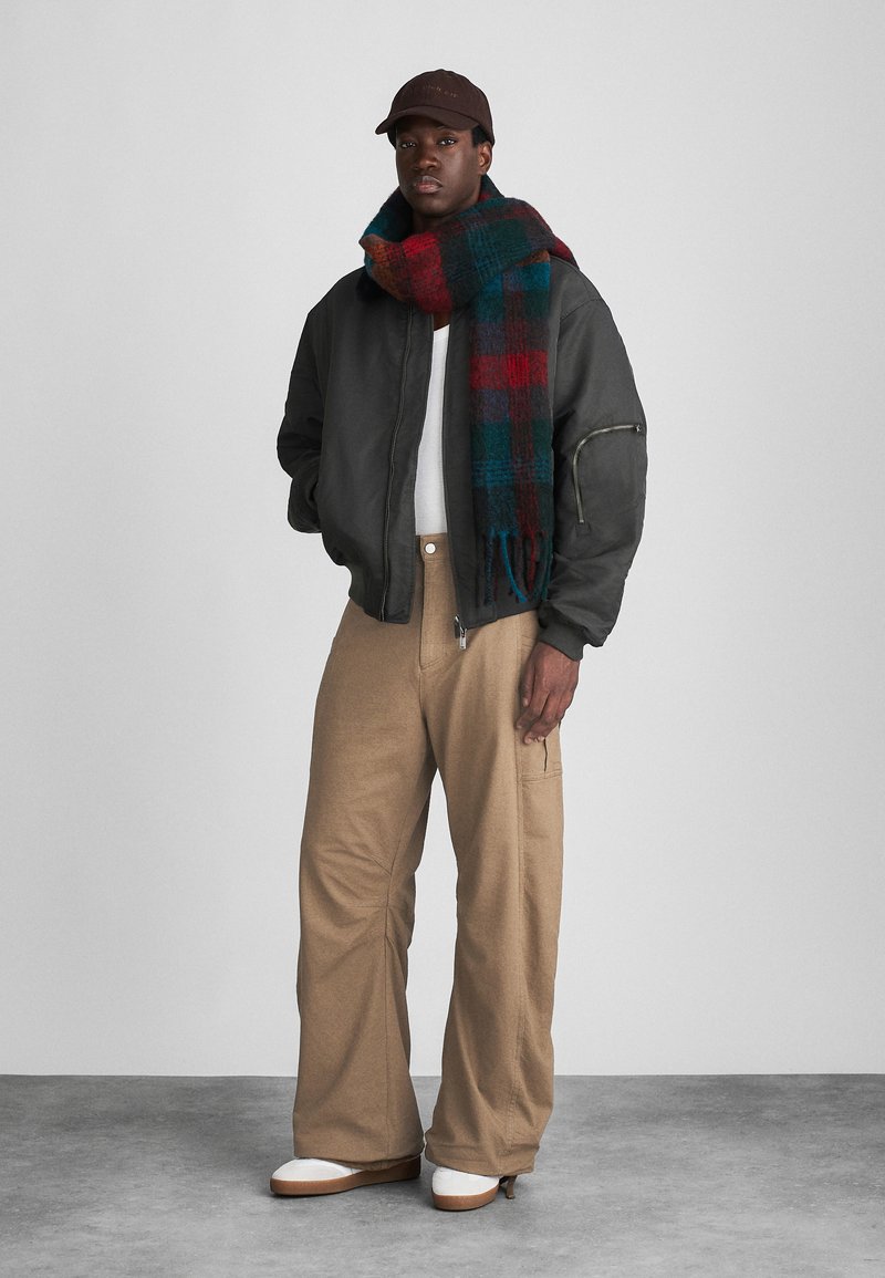 Veste bombardier noire, pull blanc, pantalons larges beiges, et écharpe à carreaux rouge, vert et bleu. Casquette marron et chaussures blanches.