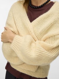 Person trägt eine grob gestrickte cremefarbene Strickjacke über einem dunkelroten Oberteil und einer schwarzen Perlenkette, die Arme vor der Brust verschränkt.