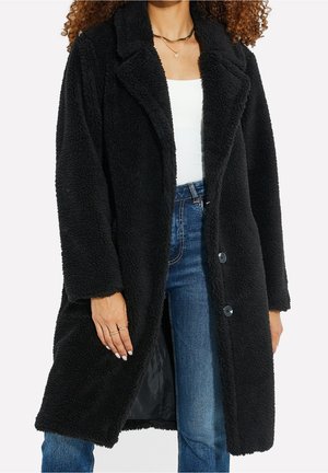 Femme portant un long manteau noir duveteux par-dessus un haut blanc et un jean bleu, montrant les détails des boutons et le tissu texturé.