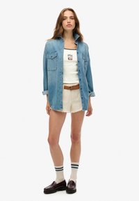 Jeanshemd, hellblau, mit Knöpfen, zwei vordere Taschen, über einem weißen Tanktop mit "USA"-Aufdruck getragen, kombiniert mit weißen, verwaschenen Shorts.