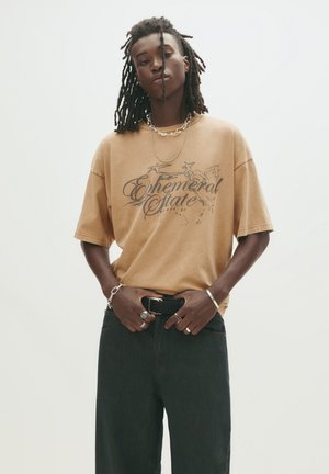 Beige oversized t-shirt med grafisk tekst, parret med mørke denim jeans. Accessories inkluderer sølv kædede halskæder og ringe.