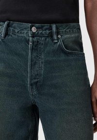Jeans in denim verde scuro, caratterizzati da una chiusura con bottone, design a cinque tasche e una leggera texture. Il tessuto presenta una delicata sfumatura e ombreggiature.