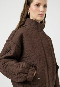 Chaqueta acolchada marrón con un patrón floral, cuello alto y puños fruncidos. Incluye bolsillos laterales y cierres de presión. Textura suave.