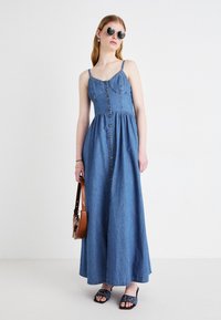 Robe maxi en denim avec un corsage ajusté, des bretelles réglables et un devant à boutons. Tissu bleu, jupe évasée, portée avec des sandales noires.