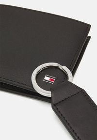 Portefeuille en cuir noir avec un anneau argenté et un logo gravé. Accent rouge, blanc et bleu marqué sur le logo. Texture lisse, design minimaliste.