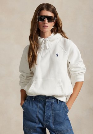 Junge Frau mit weißem Hoodie und blauen Jeans, Hände in den Taschen, und große dunkle Sonnenbrille vor neutralem Hintergrund.