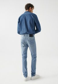 Svijetloplave traperice uskog kroja, s crnom kožnom oznakom na stražnjem pojasu i dva stražnja džepa. Uparene s plavom košuljom.