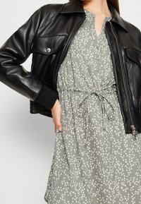 Femme portant une veste en cuir noir par-dessus une robe florale gris clair avec des boutons et une ceinture à la taille, la main sur la hanche.