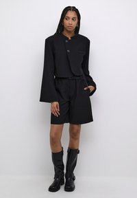 Chaqueta negra recortada con cuello redondo y dos botones, combinada con pantalones cortos negros sueltos y botas de goma negras hasta la rodilla.