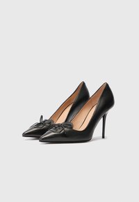 Černé stiletto podpatky s špičatou špičkou a dekorativními šňůrkami na přední části, s béžovou podšívkou, vystavené na bílém pozadí.