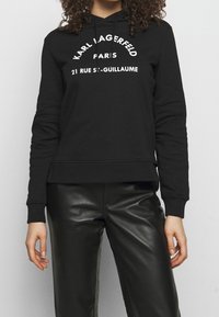 Mujer vestida con una sudadera negra con el texto "KARL LAGERFELD PARIS 21 RUE ST-GUILLAUME" y pantalones de cuero negros, de pie contra un fondo liso.