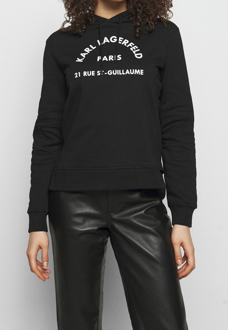Mujer vestida con una sudadera negra con el texto "KARL LAGERFELD PARIS 21 RUE ST-GUILLAUME" y pantalones de cuero negros, de pie contra un fondo liso.