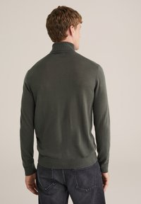 Mörkgrön stickad tröja med hög krage, långa ärmar och ribbade muddar. Slät textur och figurnära design, bärs över mörka jeans.