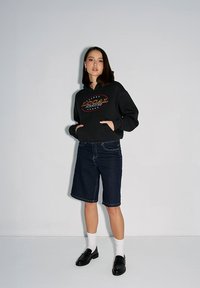 Czarna bluza z kapturem z grafiką "LOCAL" w czerwonym i białym kolorze, ciemnoniebieskie bermudy, białe skarpety i czarne mokasyny. Gładkie tekstury, casualowy design.