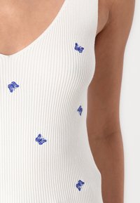 Geribbelde witte tanktop met verspreide blauwe vlinderborduring. De halslijn heeft een diepe V-vorm, wat de gestructureerde stof benadrukt.