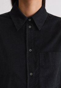 Chemise en velours côtelé noire avec un col à revers, une fermeture boutonnée à l'avant et une poche poitrine. Caractérisée par un tissu texturé et des boutons gris.