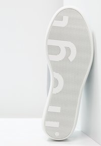 Witte sneaker met een grijze textielen bovenkant, een gestructureerde rubberen zool met horizontale groeven en grote gegraveerde letters, gezien van onderaf.